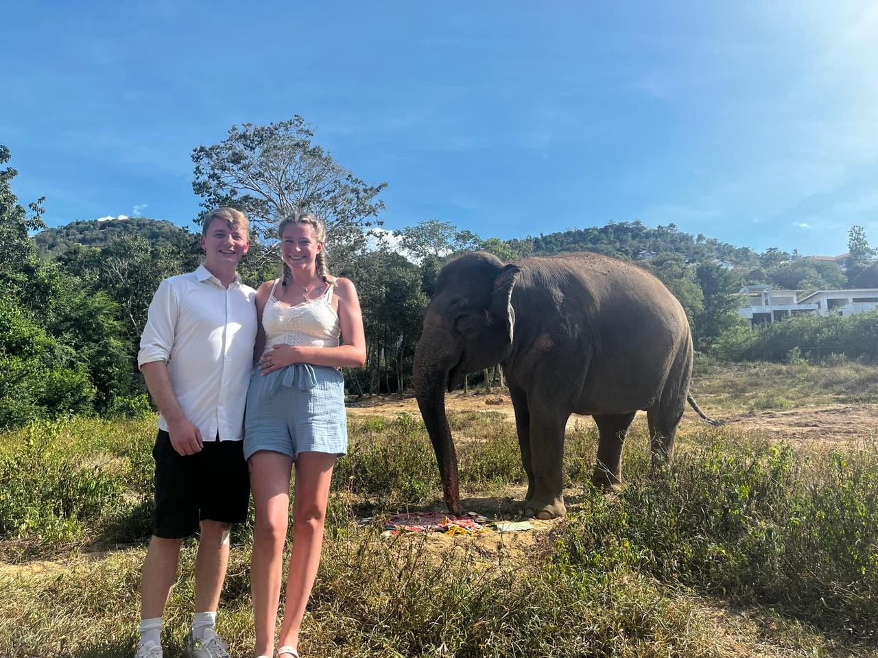 Shawn and Kate standing together outdoors in Koh Samui, with an elephant beside them and forested hills behind.